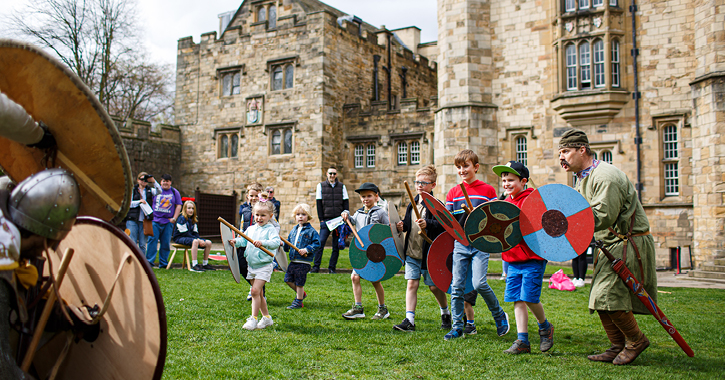 group of children holding swords and shields play fighting with medieval reenactors at Durham Castle.